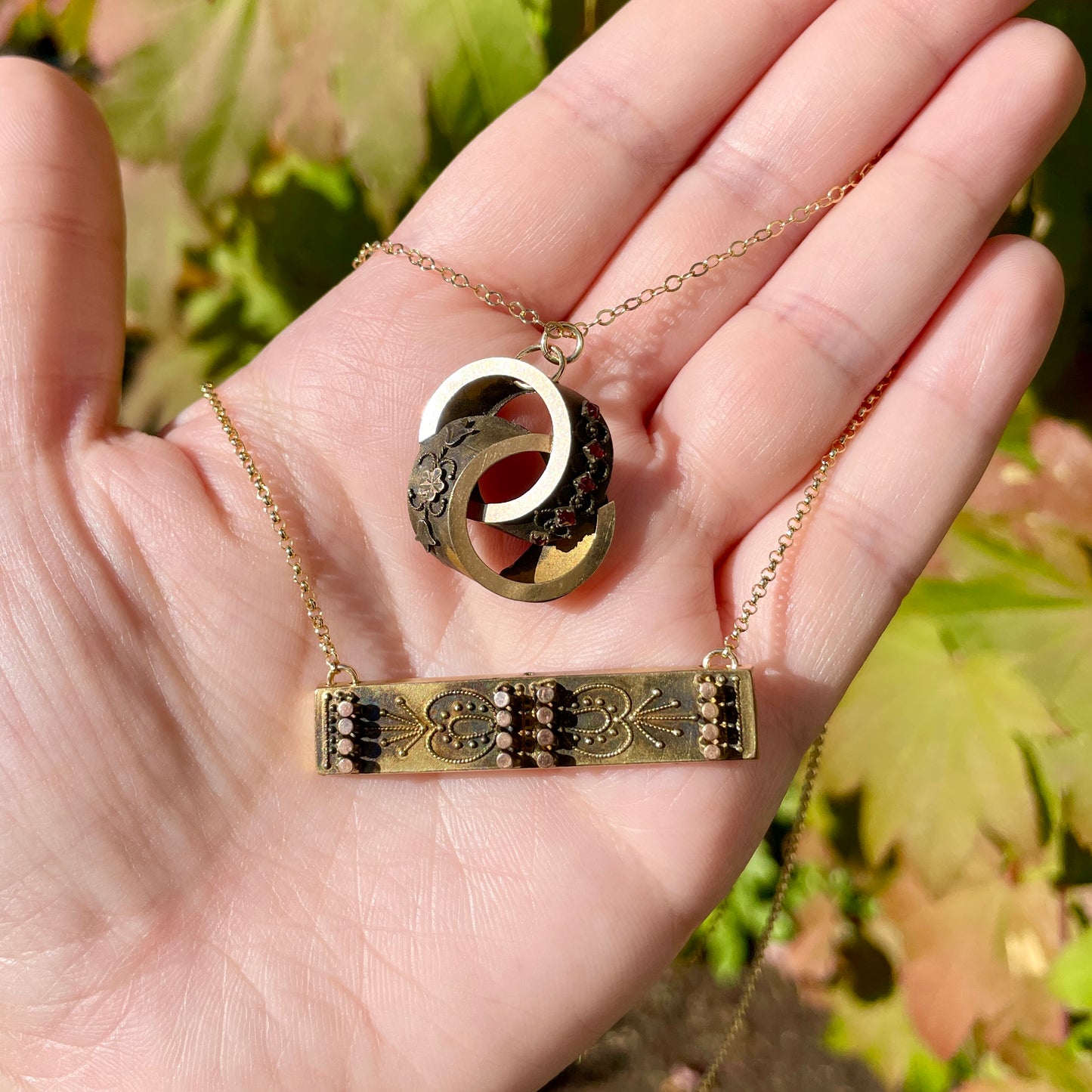 Two Etruscan revival bar pin and brooch pin conversion necklaces with 14k gold filled chain, being held in the palm of hand in the sunlight.