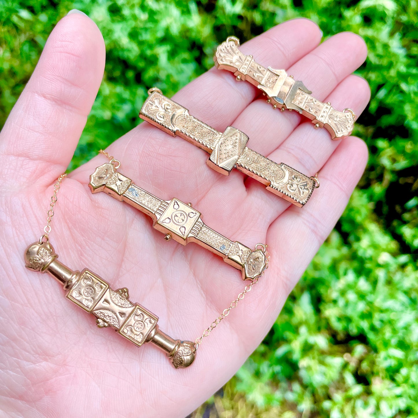 Four antique Victorian bar pin necklaces laying across the palm and fingers of a left hand with green shrubbery in the background.
