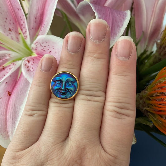 Hand wearing a ring with a face design in front of pink flowers