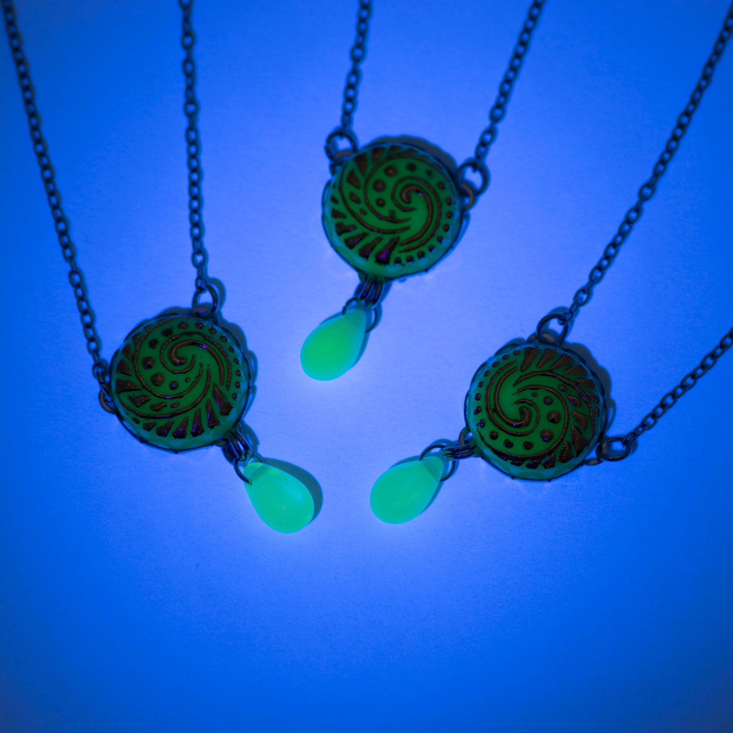 Three button pendant necklaces glowing from a UV blacklight. Uranium glass button in light turquoise color.  Uranium glass button contains 2% uranium dioxide. This small amount in the glass allows it to fluoresce (glow) green under a black light and is also safe to wear.