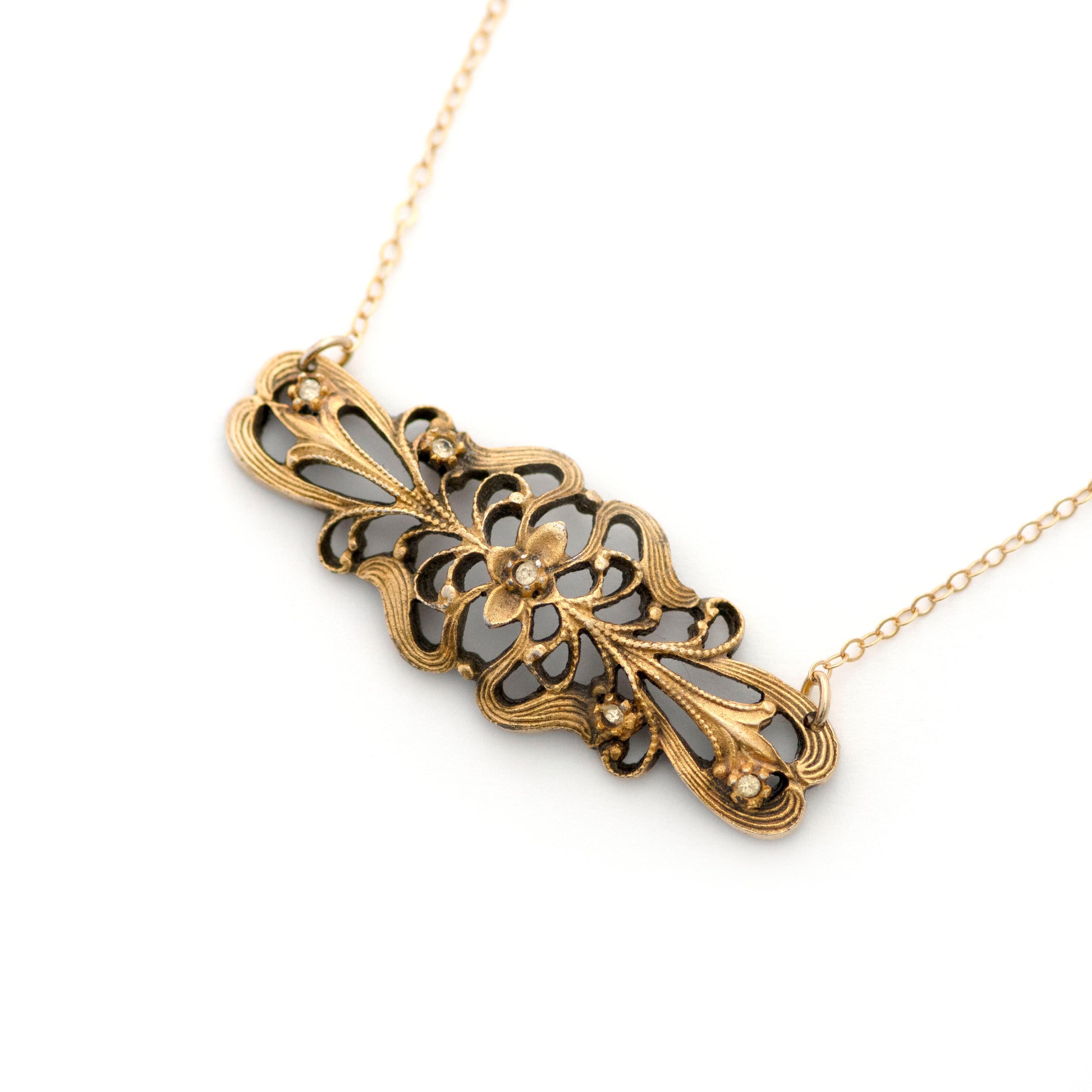 A product photography image of a vintage bar pin repurposed pendant dangling on a 14k gold filled cable chain on a white background. Pendant has a scrolling floral swirls in the style of Art Nouveau era design and is studded with 5 clear paste rhinestones.