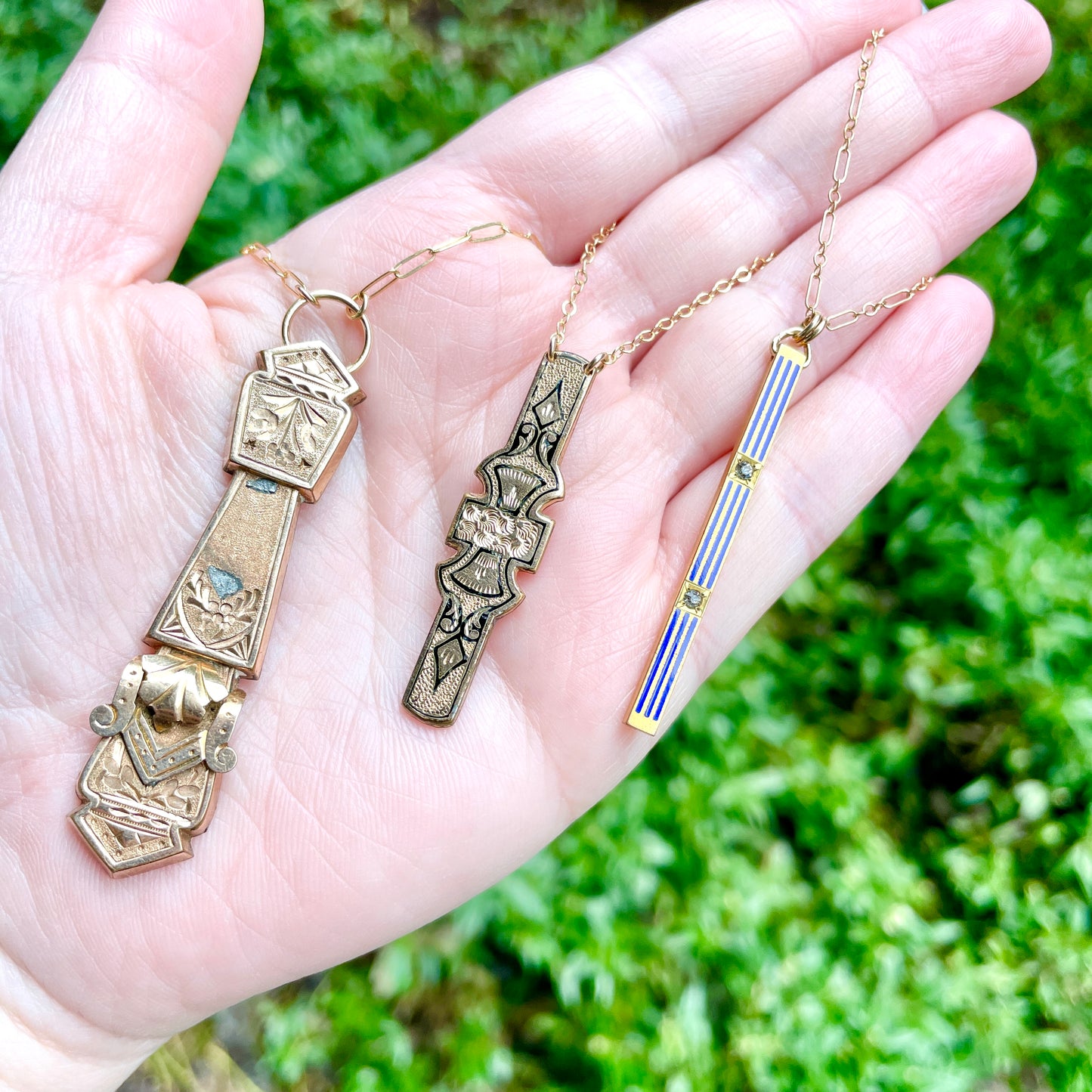 Three antique vertical bar pin pendant necklaces held on a flat palm of a left hand with green foliage in the background.
