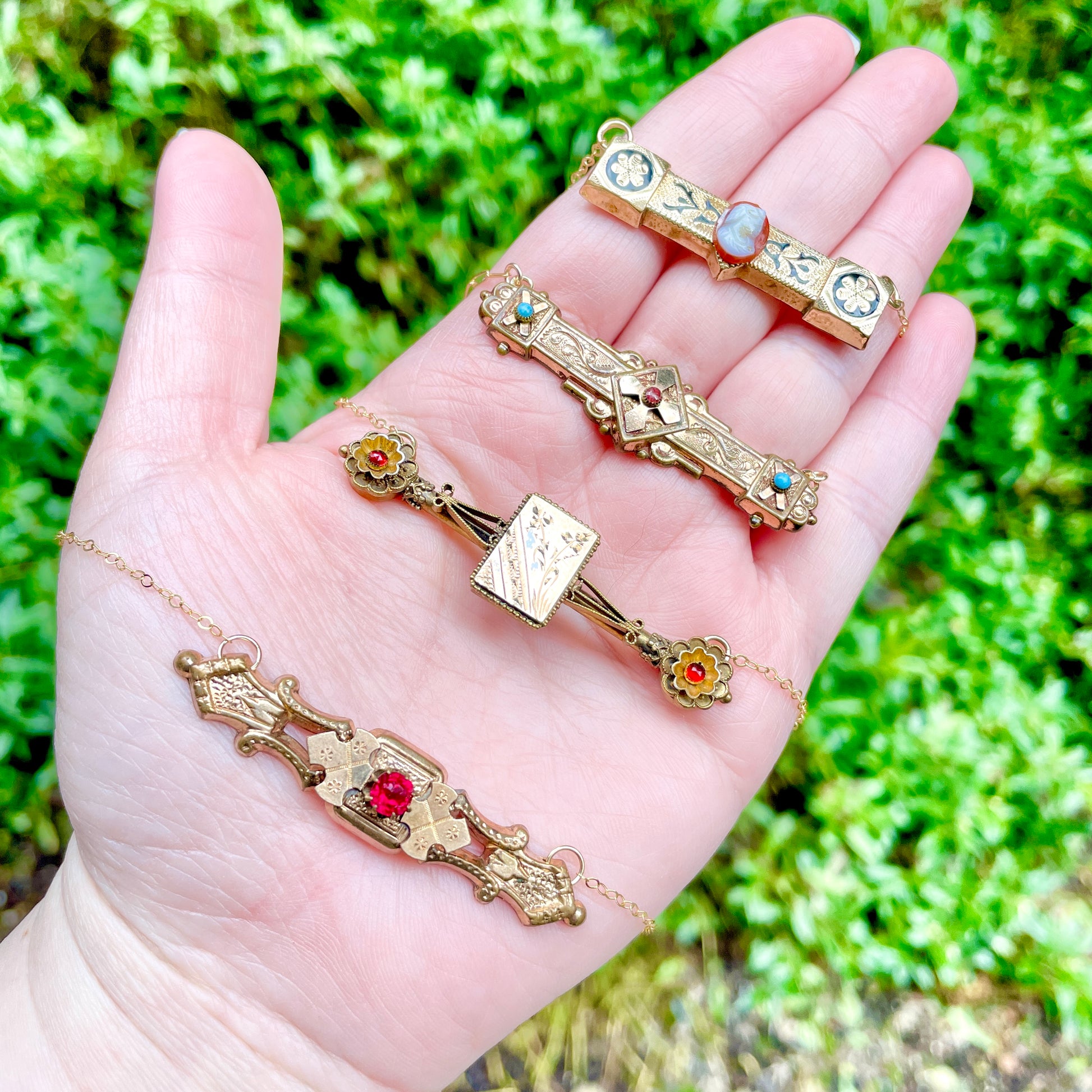 these are four victorian bar pin necklaces laying across the palm side of a left hand with greenery in the background