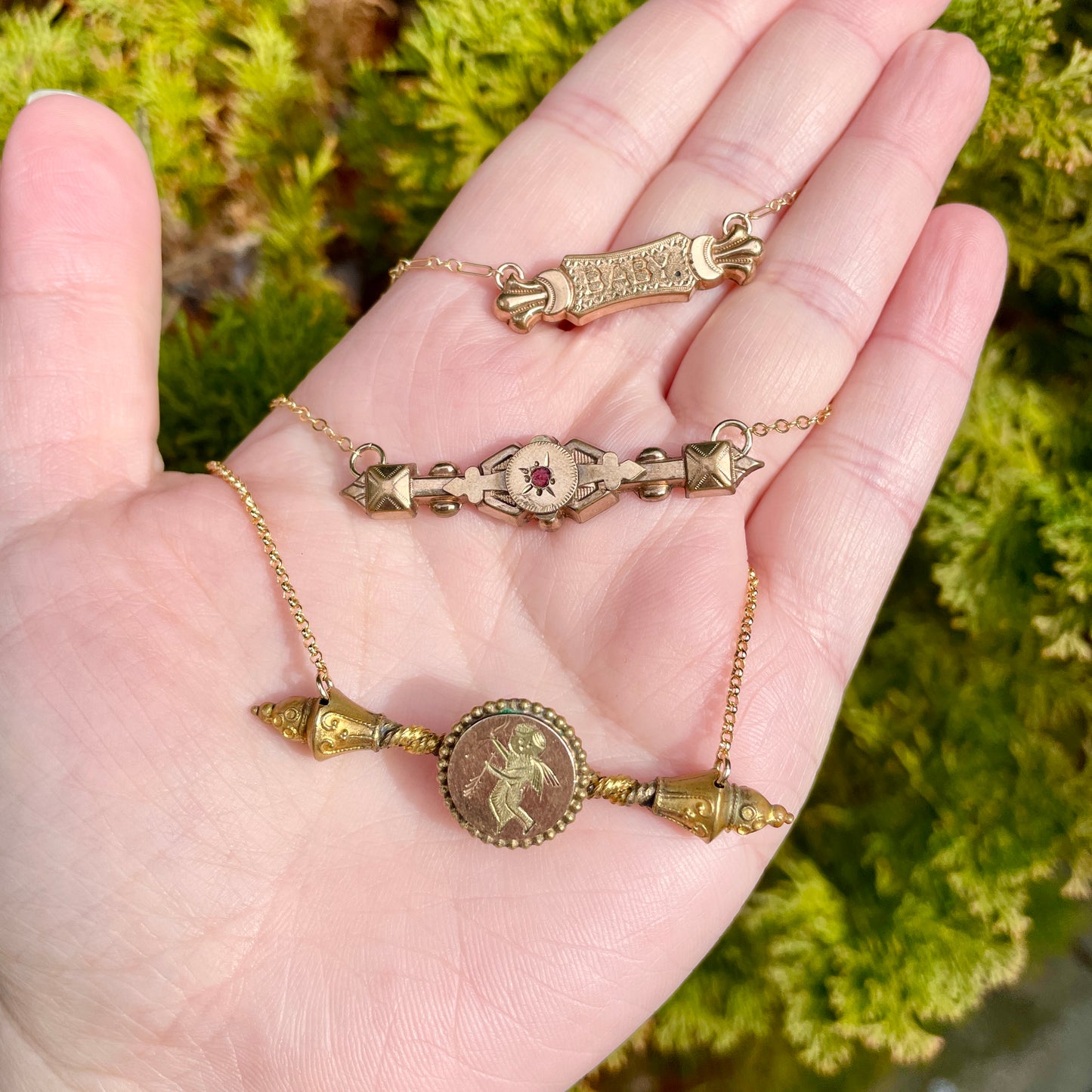 3 bar pin necklaces in palm of hand on background of evergreen foliage: Baby bar pin necklace, red paste edwardian bar pin necklace, cupid with his bow and arrow bar pin necklace