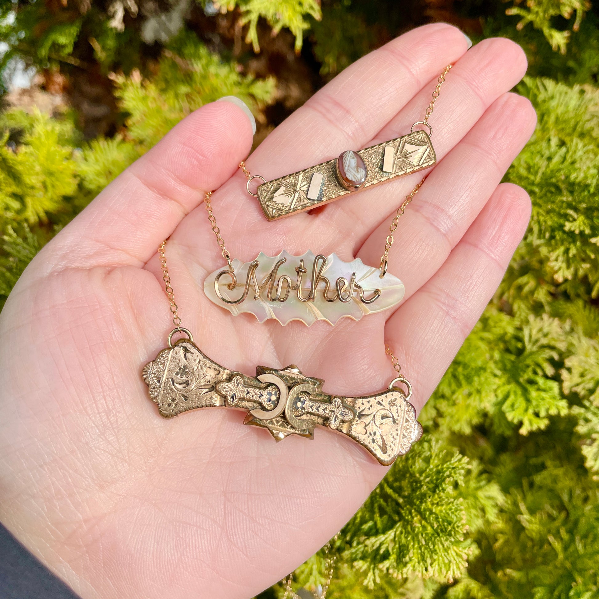 3 vintage and antique bar pin necklaces held on palm side of hand with green foliage background.