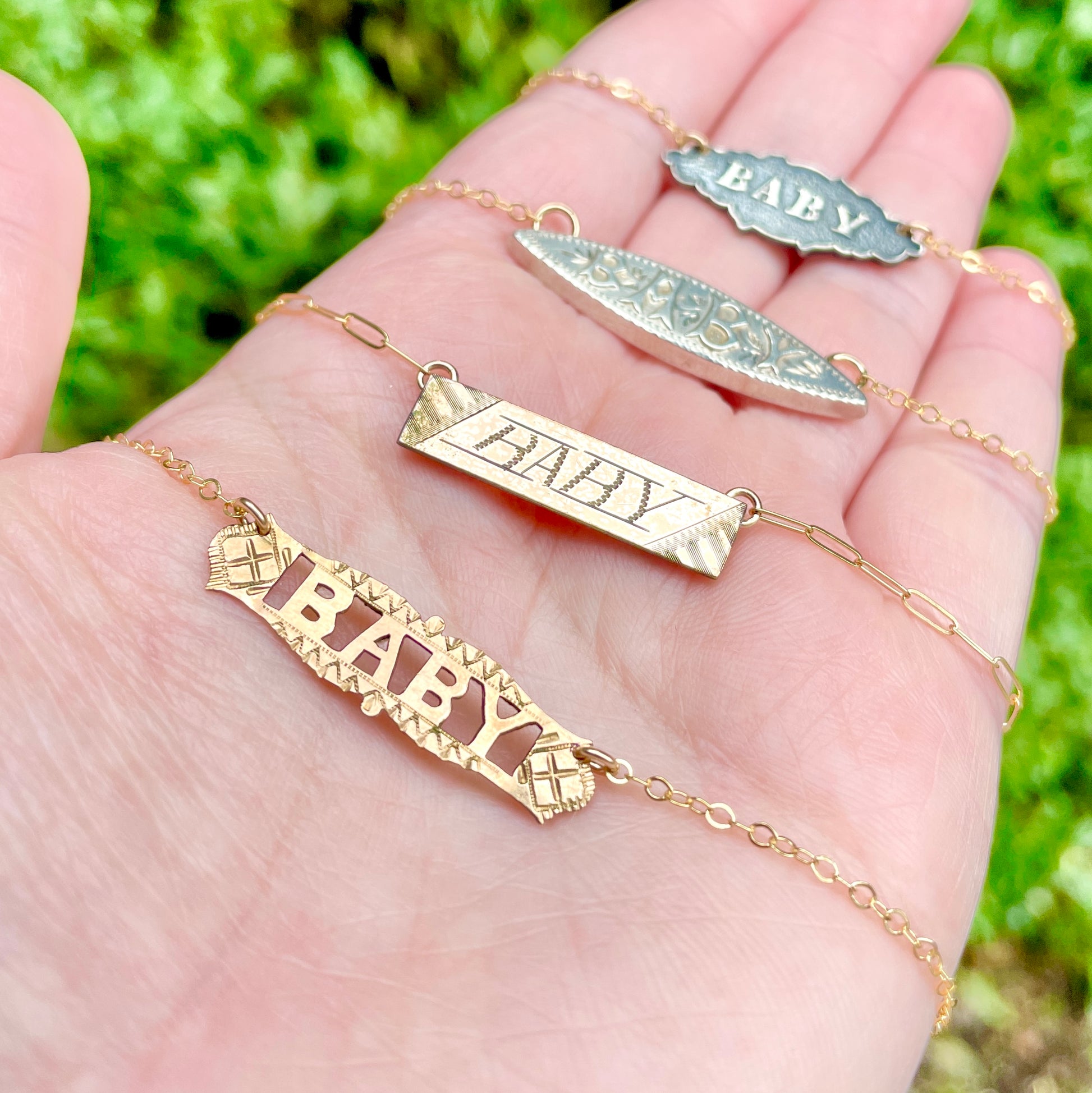Four bar pin and brooch conversion necklaces in gold filled and fine silver, each depicting the word BABY. Necklace are shown on the palm side of the left hand with greenery in the background.