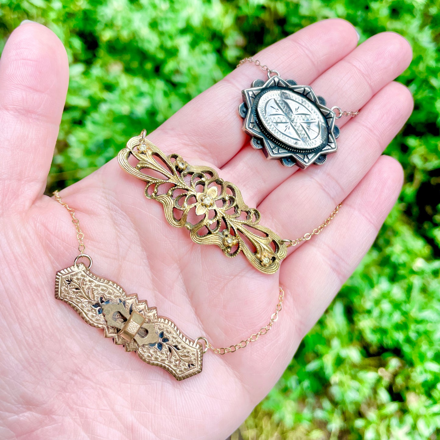 Three repurposed vintage brooch pendants laying on the palm side of a hand with green foliage in the background.