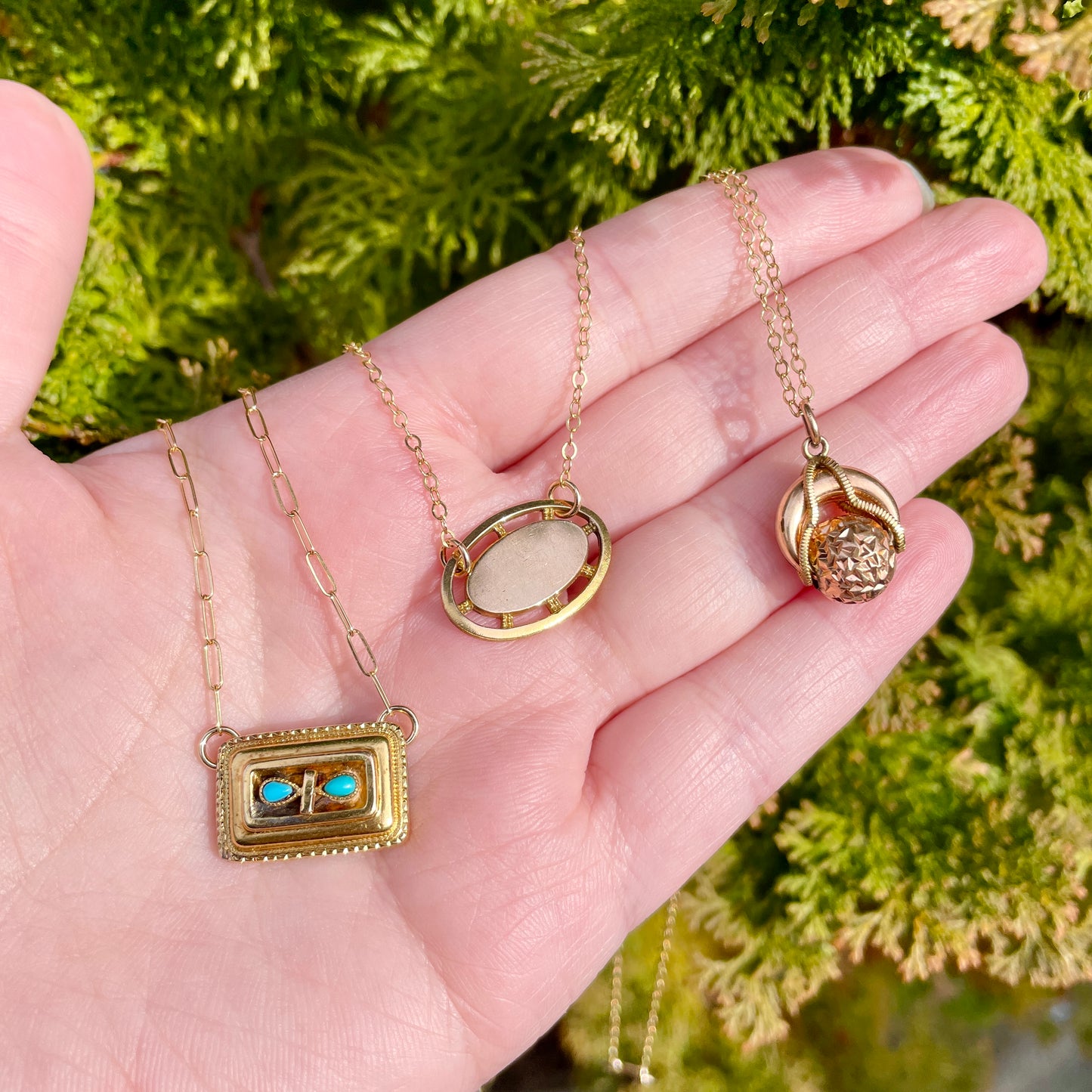 three small antique pendant necklaces held on the palm side of a hand against a green natural foliage background