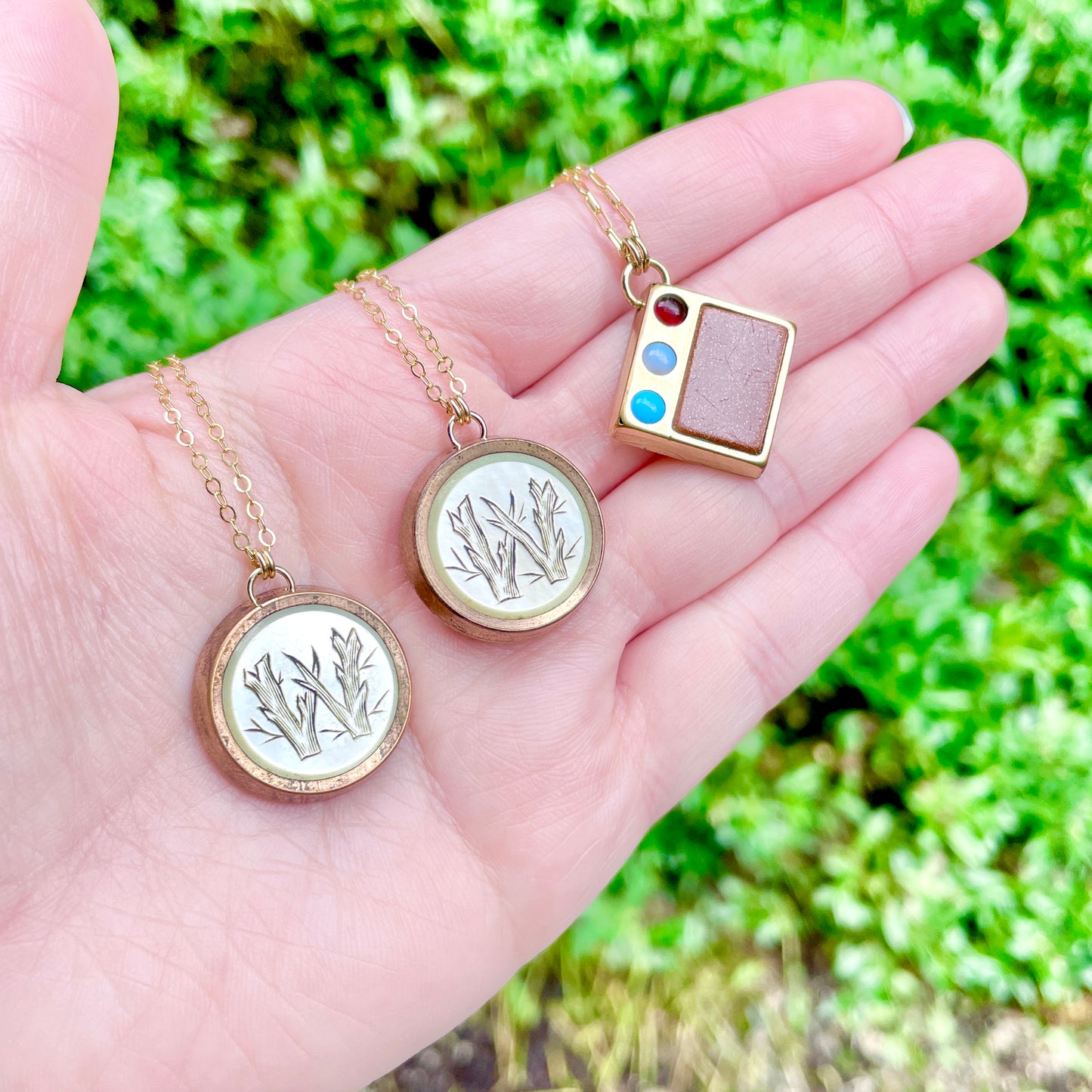 Three antique cufflink conversion pendant necklaces laying on the pal side of a left hand with green foliage in the background.