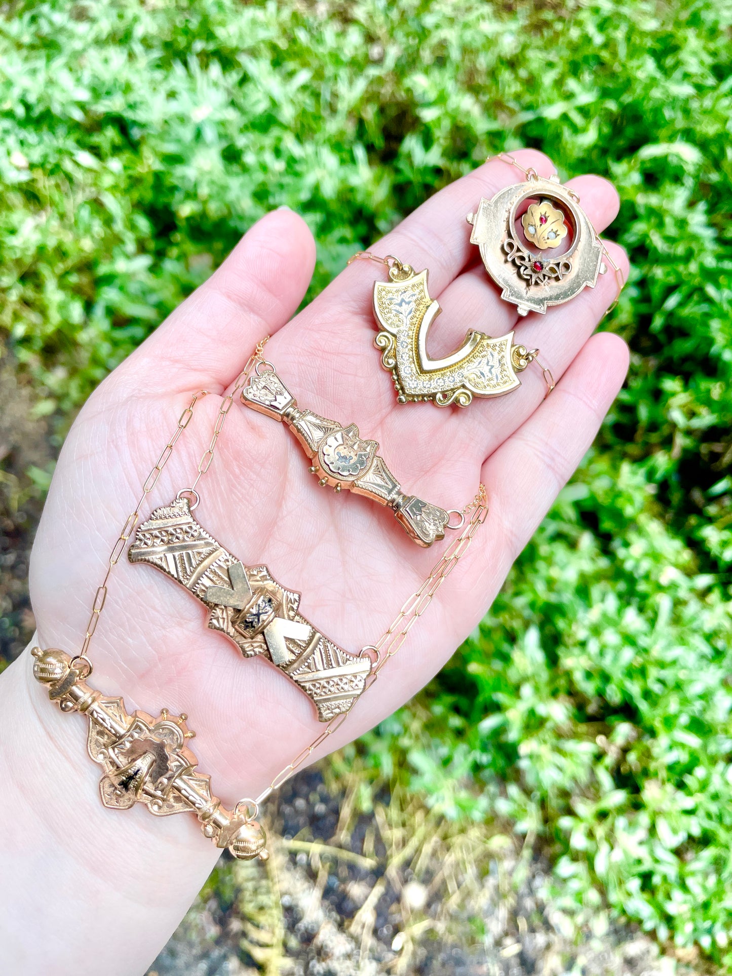 five brooch and bar pin conversion necklaces displayed on palm side of a left hand hovering over a green foliage background